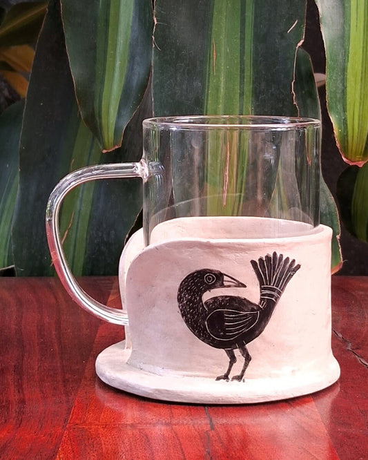 A clear borosilicate mug placed on top of a white papier-mache mug holder with a hand-painted black bird design.