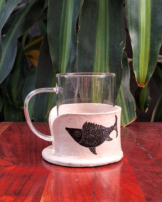 A clear glass mug with a beige papier-mache holder featuring a black fish design, placed on a wooden surface with green leafy background.