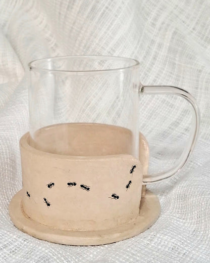 A clear glass mug placed on top of a beige papier-mache mug holder with black ant designs.
