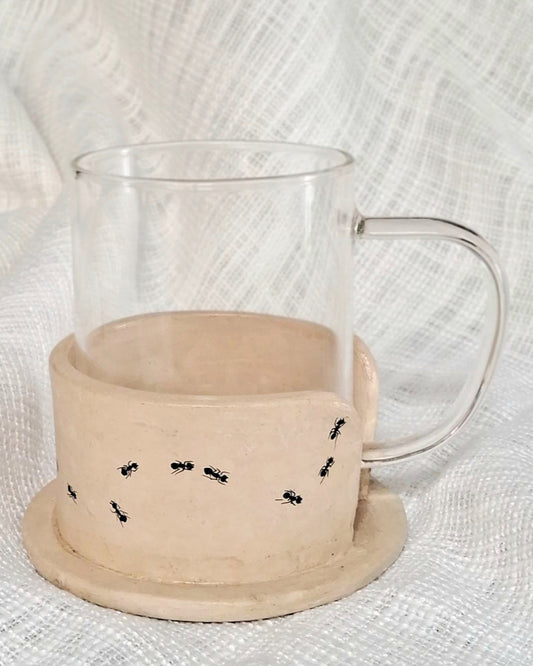 A clear glass mug placed on top of a beige papier-mache mug holder with black ant designs.